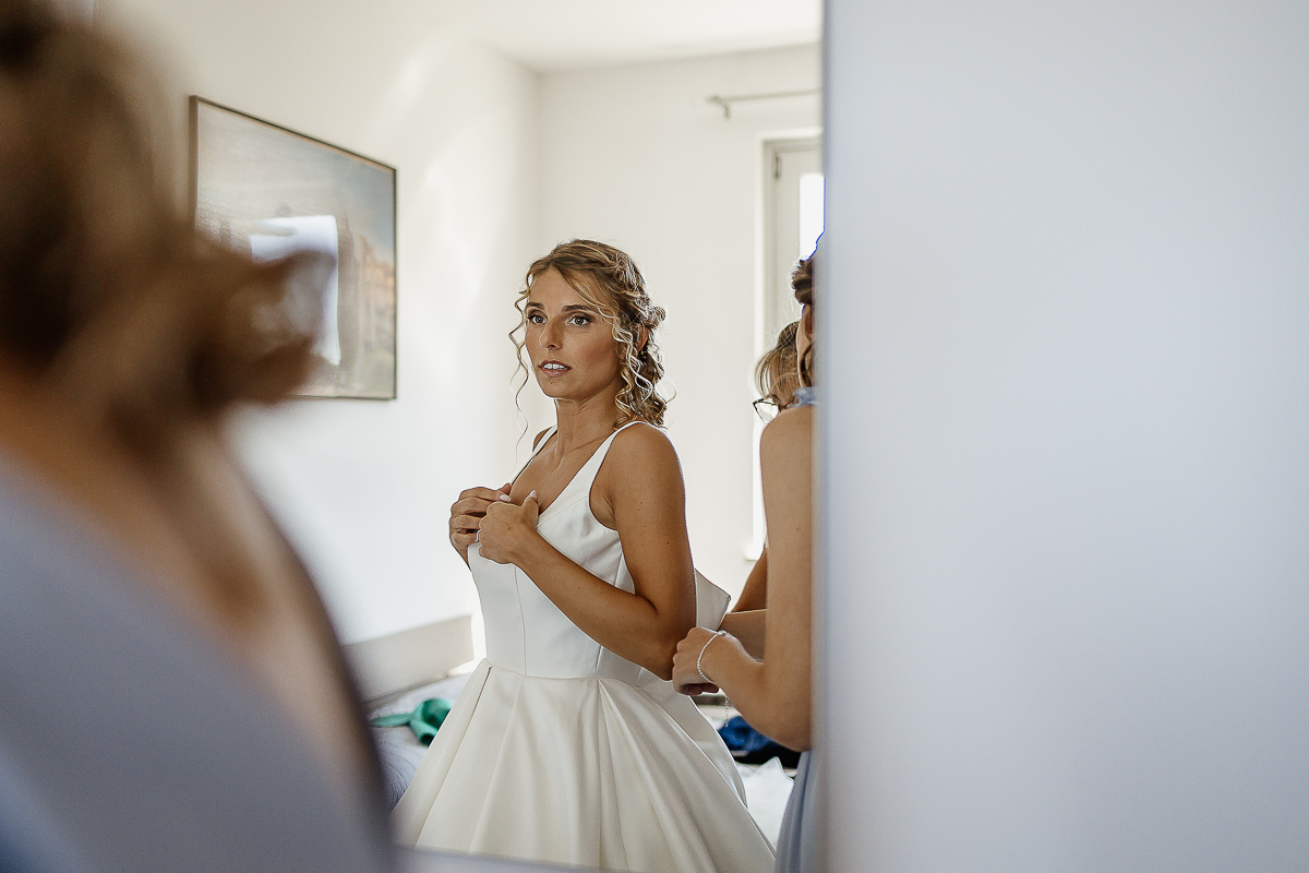 matrimonio a Chieri, preparazione della sposa, sposa allo specchio mentre la mamma e la testimone le abbottonano l'abito