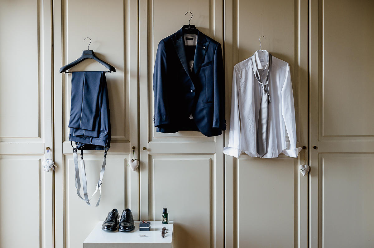 matrimonio a Chieri, preparazione dello sposo, pantaloni, giacca e camicia appesi all'armadio bianco, accessori sul comodino