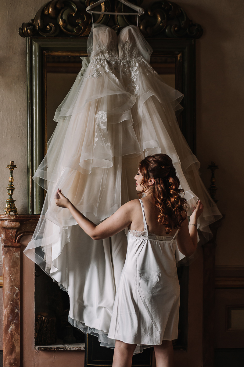 Matrimonio al Castello Canalis di Cumiana(TO). Sposa che accarezza il suo bellissimo abito da sposa appeso alla specchiera sul camino