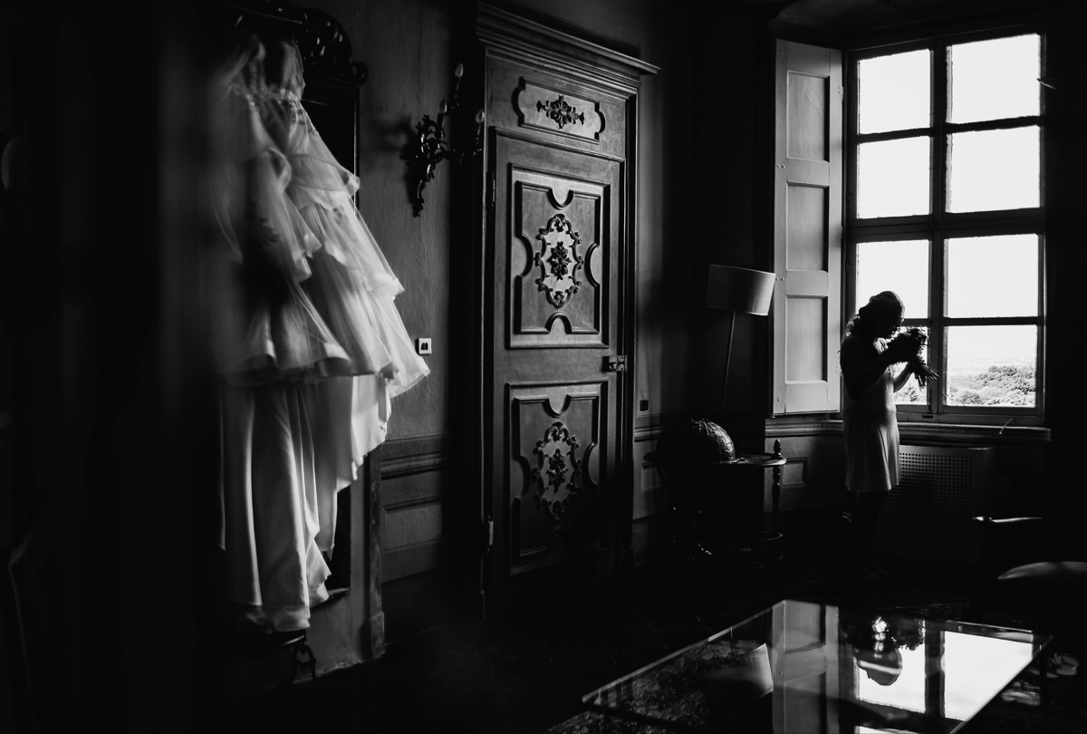 Matrimonio al Castello Canalis di Cumiana(TO). Scena ripresa dallo specchio di un mobile trumeau settecentesco. Sposa alla finestra con il bouqut. abito appeso alla specchiera del camino. Bianco e nero.