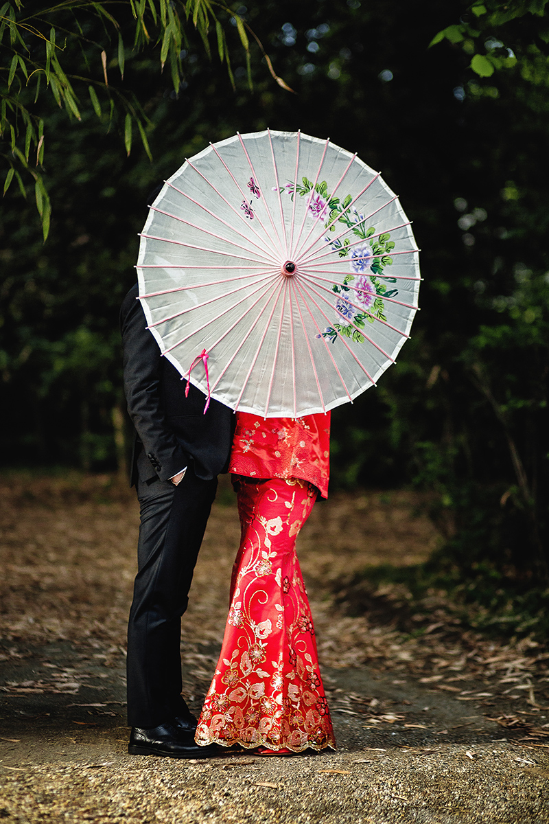 Castello di Collegno, matrimonio cinese. Cerimonia tradizionale. Sessione fotografica di coppia. sposa indossa una giacca tradizionale cinese di seta rossa ricamata.Sposi camminano nel bosco dei bambù. Ritratto degli sposi dietro l'ombrellino di carta di riso tradizionale cinese.