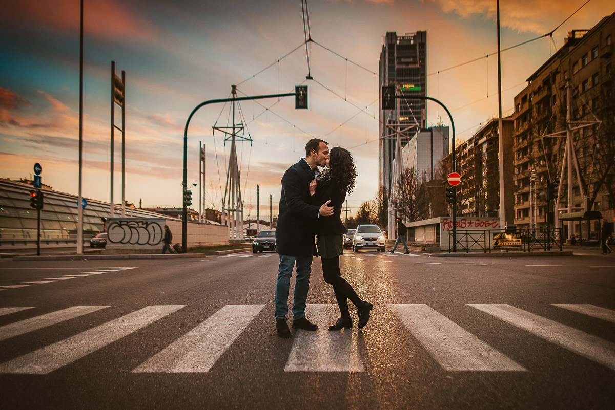 Engagement a Torino nella stazione di Porta Susa. Coppia al tramono, sulle strisce pedonali di corso Inghilterra davanti al grattacielo Intesa San Paolo. le auto dietro con i fari accesi.