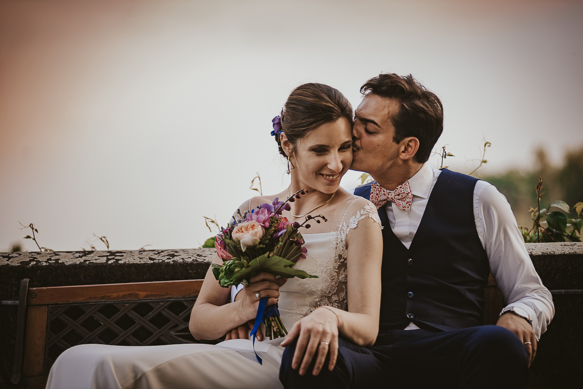 Castello Rosso di Costigliole di Saluzzo. Destination wedding. Sessione di coppia al tramonto. Sposi seduti, sposo bacia la sposa sulla guancia.