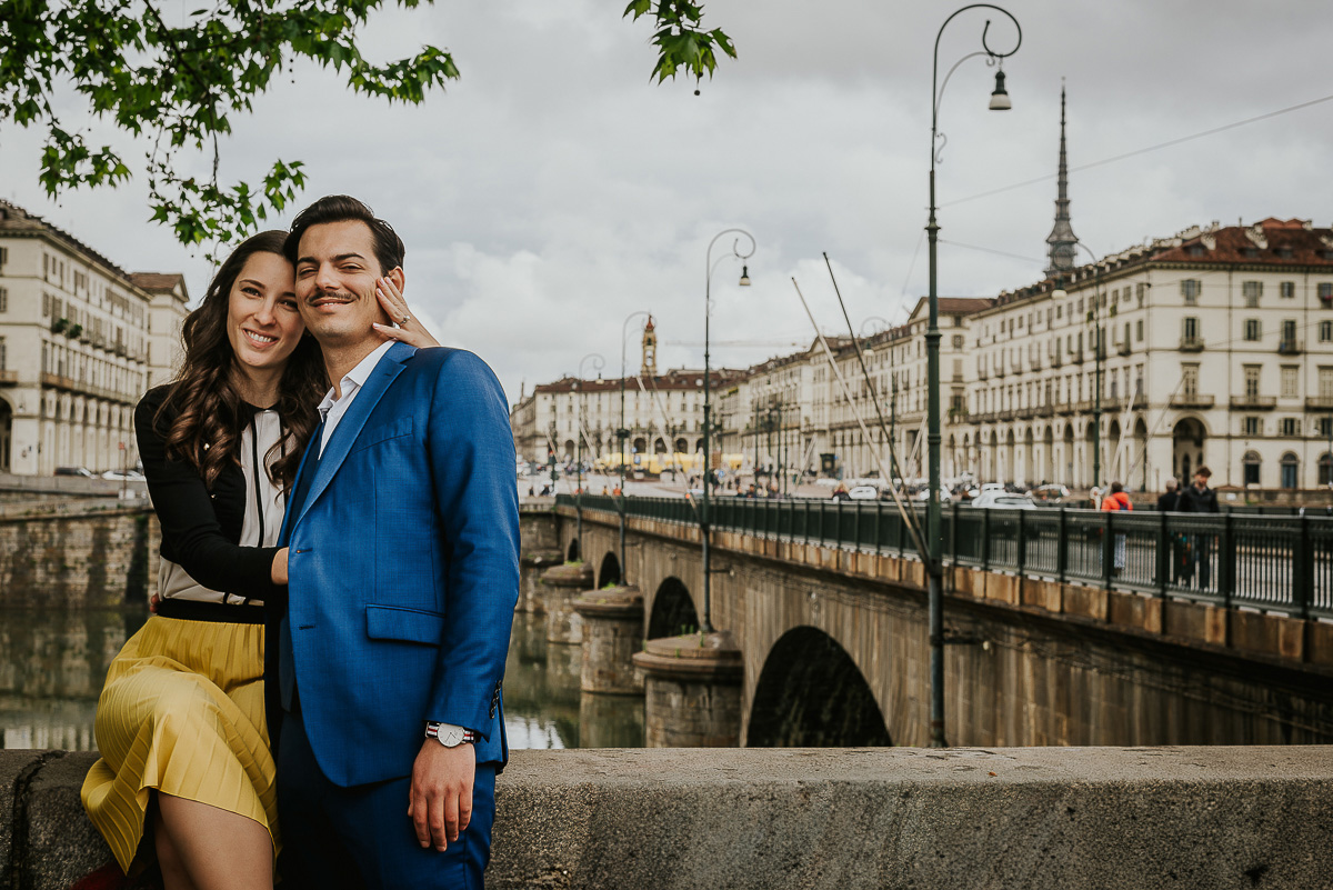 Engagement a Torino, in piazza Vittorio Veneto a Torino di Sara e Gabriel. Coppia seduta sul muretto degli argini del Po con la piazza sullo sfondo.