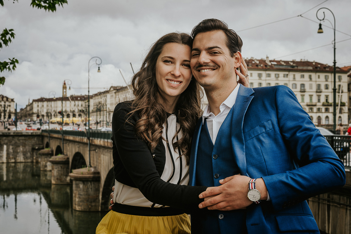 Engagement a Torino, in piazza Vittorio Veneto a Torino di Sara e Gabriel. Coppia seduta sul muretto degli argini del Po con la piazza sullo sfondo.