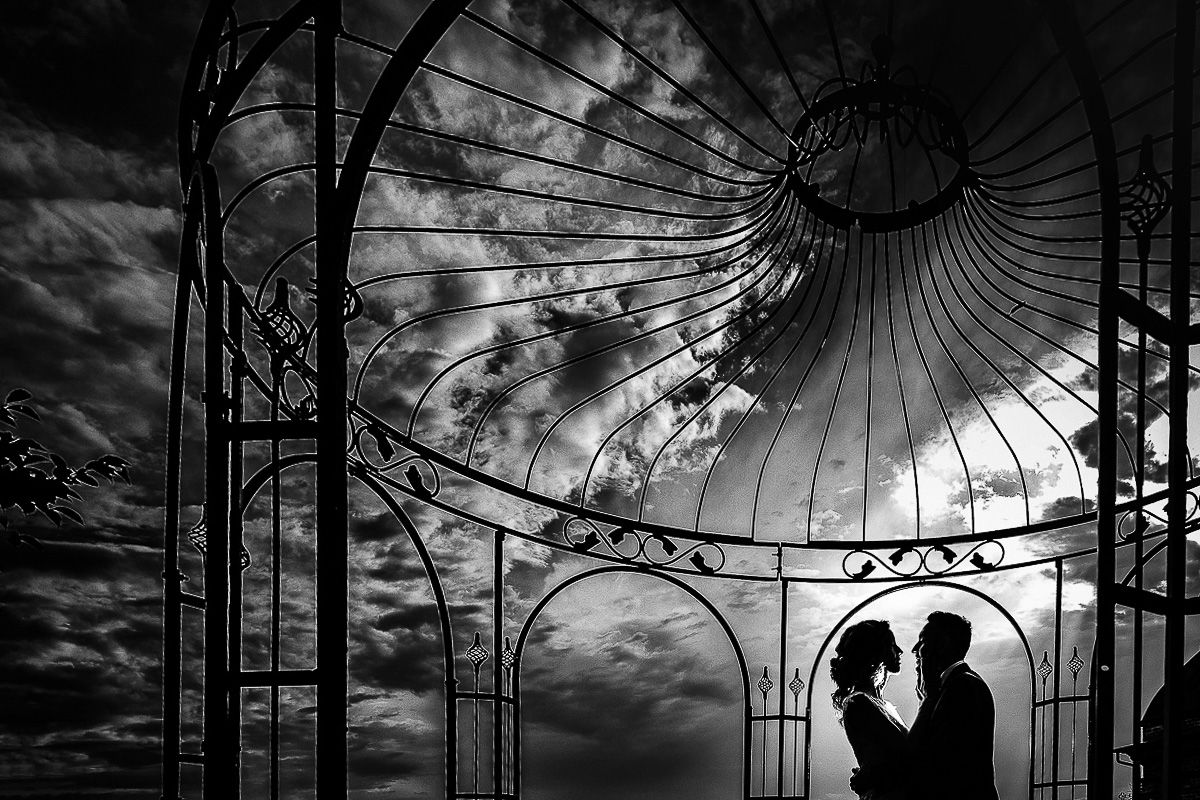 Matrimonio al castello di Cortanze. Ricevimento. Sessione di coppia. silhouette degli sposi in bianco e nero. Sotto un gazebo in ferro battuto con cielo drammatico gonfio di nubi.