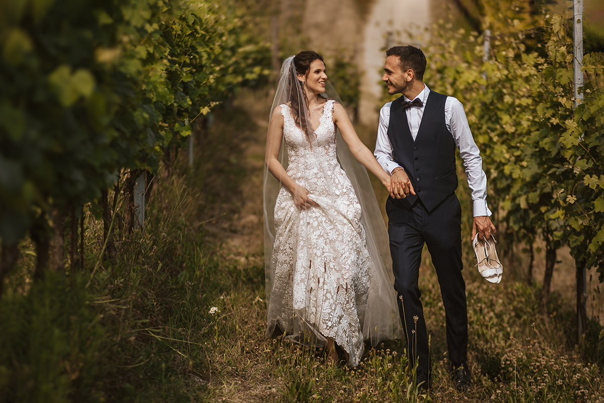 Tenuta Fontanfredda, Guidoristorante. Villa reale.Sessione fotografica degli sposi tra le vigne. Sposiche passeggiano. Sposo tiene in mano le scarpe della sposa.
