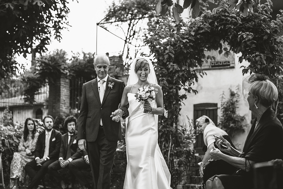 Matrimonio a Villa Delfini di Morsasco (AL) nel Monferrato. Cerimonia. sposa al braccio del padre, saluta ridendo un'invitata.
