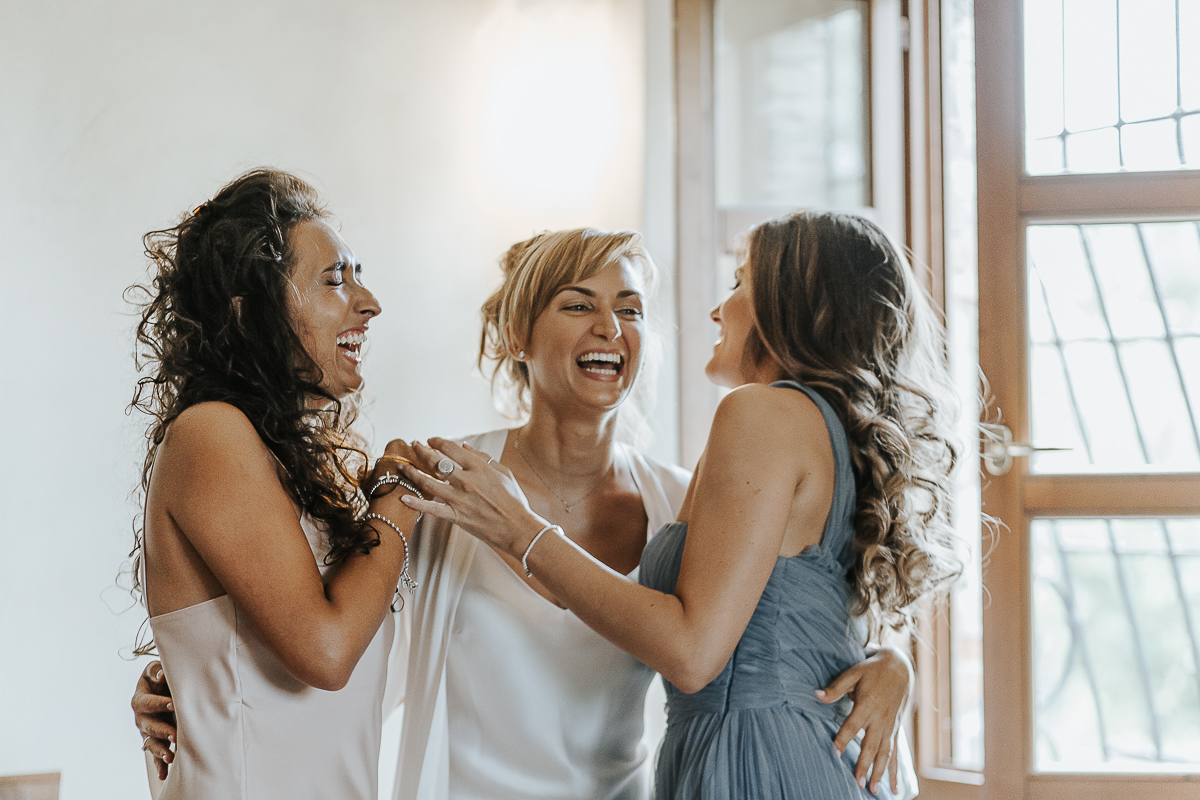 Matrimonio a Villa Delfini di Morsasco (AL) nel Monferrato. Preparazione dell a sposa. Sposa abbraccia ridendo damigella d'onore e testimone.