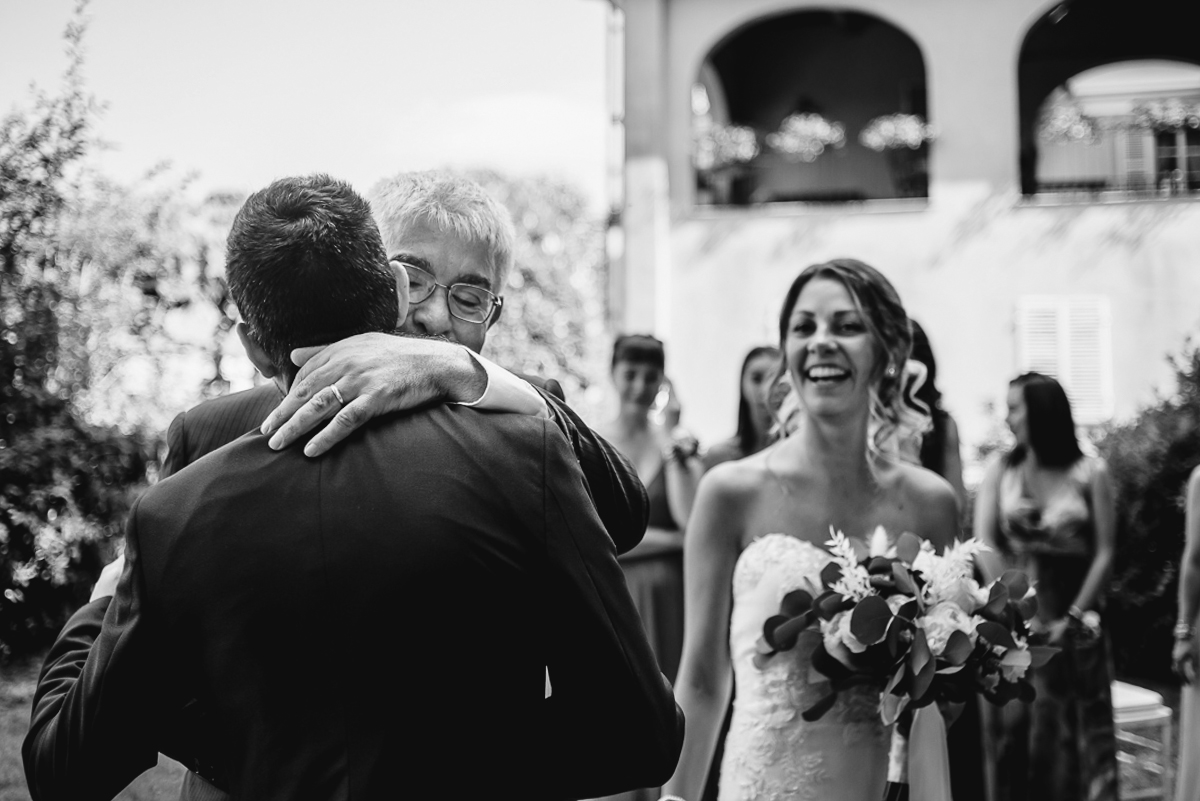 Matrimonio a Villa Fiorita ad Asti. Cerimonia.Padre della sposa che abbraccia lo sposo e sposa che ride.