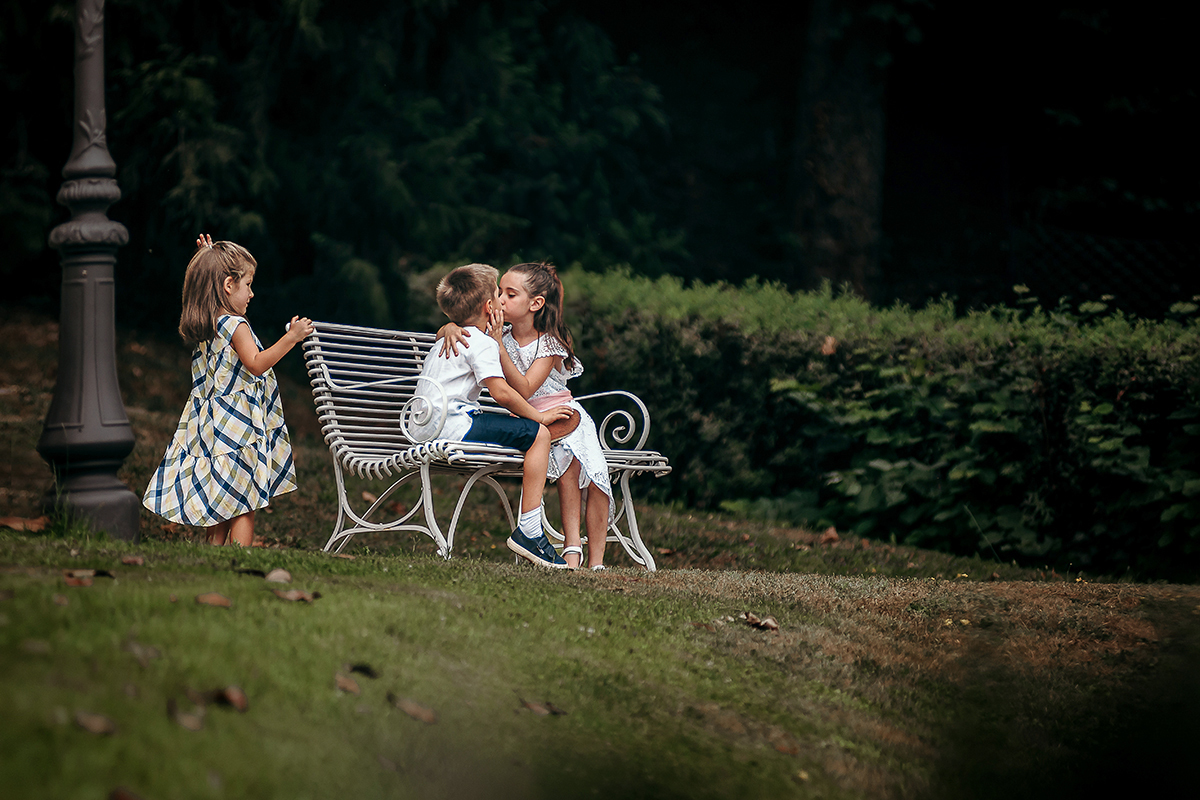 Villa Sassi matrimonio di Giulia e Nicolò. Aperitivo. bambini che si baciano seduti sui di una panchina di ferro battuto bianco, con un'altra bambina che li guarda.