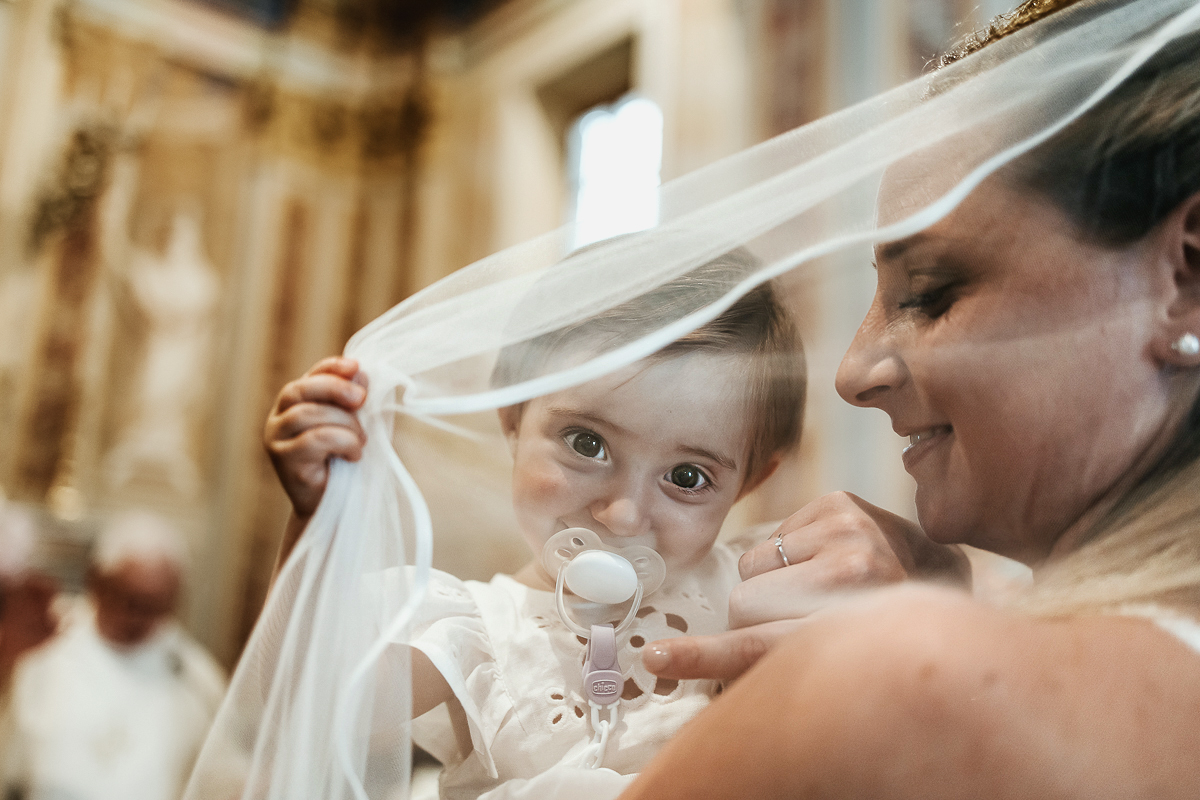 Villa Sassi matrimonio di Giulia e Nicolò. Cerimonia nella chiesa di santa Maria della Spina a Baldissero Torinese, rito del Battesimo. Bambina gioca con il velo della mamma.