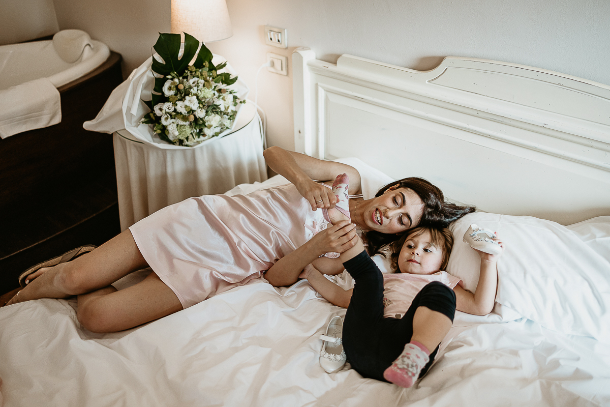 Tenuta La Cascinetta di Buriasco. Matrimonio. Preparazione della sposa. Sposa sdraiata sul letto con la bambina.