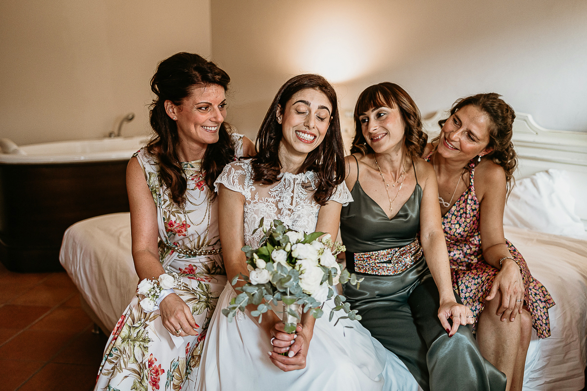 Tenuta La Cascinetta di Buriasco. Matrimonio. Preparazione della sposa. Sposa con una buffa espressione seduta sul letto con le amiche.