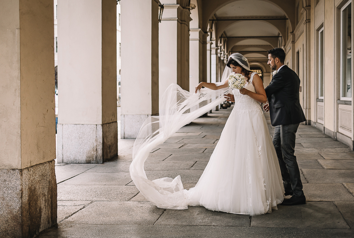 Matrimonio a Torino. Sessione di coppia davanti alla stazione di Porta Nuova. Sposa si sistema il velo sotto i portici di via Sacchi.