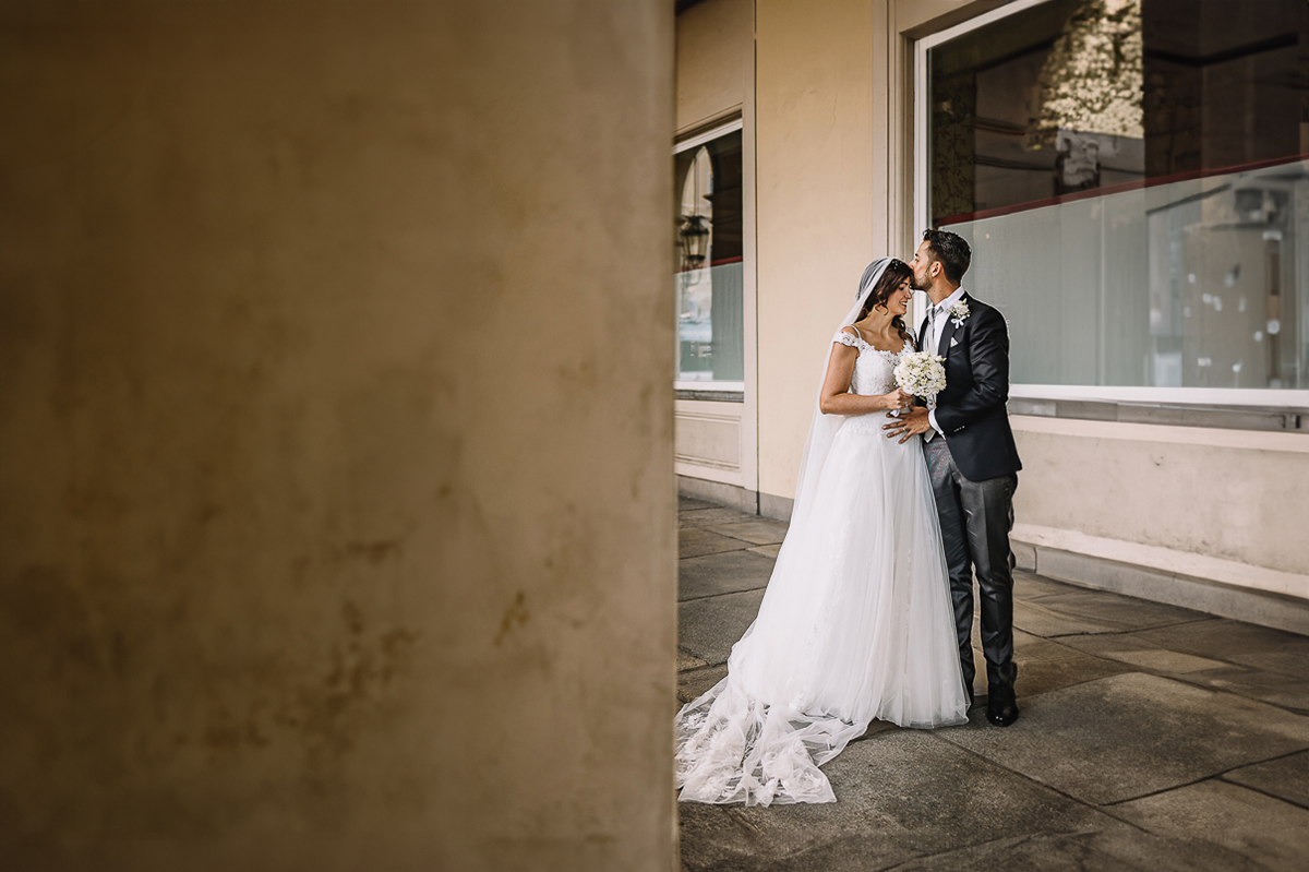 Matrimonio a Torino. Sessione di coppia davanti alla stazione di Porta Nuova. Sposo bacia la sposa sulla fronte sotto i portici di via Sacchi.