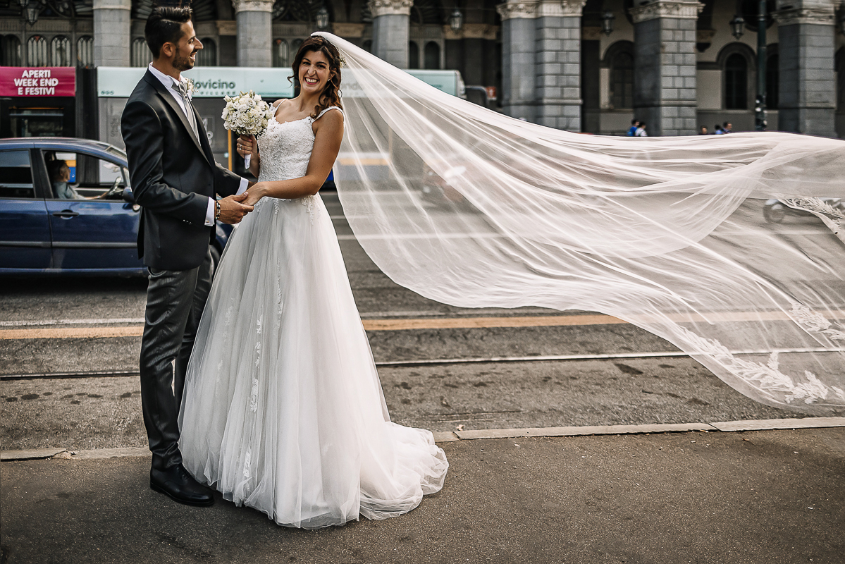 Matrimonio a Torino. Sessione di coppia davanti alla stazione di Porta Nuova. Velo sollevato da una passante.