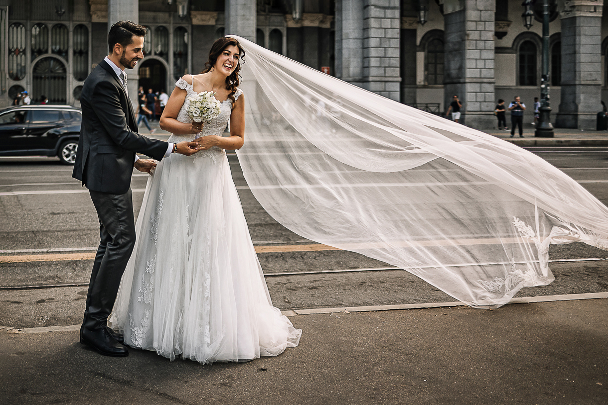 Matrimonio a Torino. Sessione di coppia davanti alla stazione di Porta Nuova. velo sollevato da una passante che quasi lo stacca dalla testa della sposa.