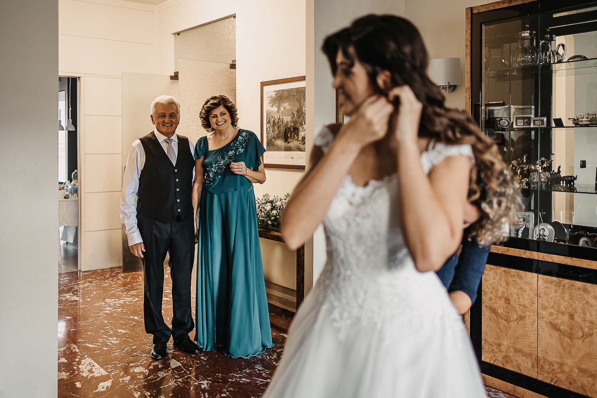 Matrimonio a Torino, preparazione della sposa. Sposa si mette gli orecchini. Genitori sullo sfondono la guardano con amore.