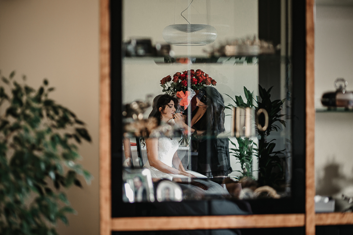 Matrimonio a Torino, preparazione della sposa. Sposa al trucco ripresa dal vetro di un mobile del soggiorno.