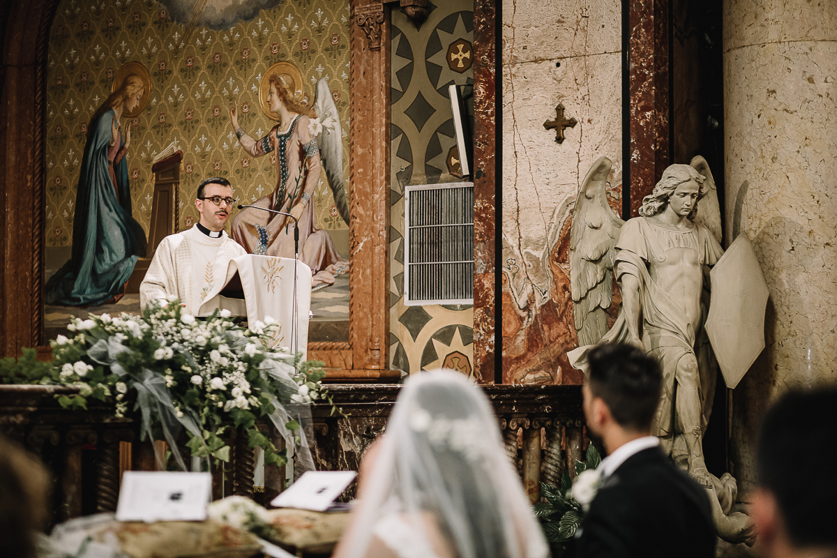 Matrimonio a Torino, cerimonia chiesa dei Santi Angeli Custodi. messa. giovane sacerdote e statua dell'angelo nella stessa posizione.