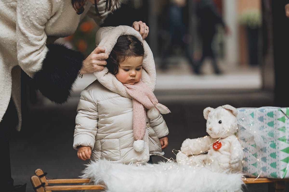 Fotografia di famiglia lifestyle di Natale. La piccola Lavinia con la sua mamma con un vecchio slittino di legno addobbato con un pellicciotto di lana di agnello bianco e pacchi regalo natalizi.