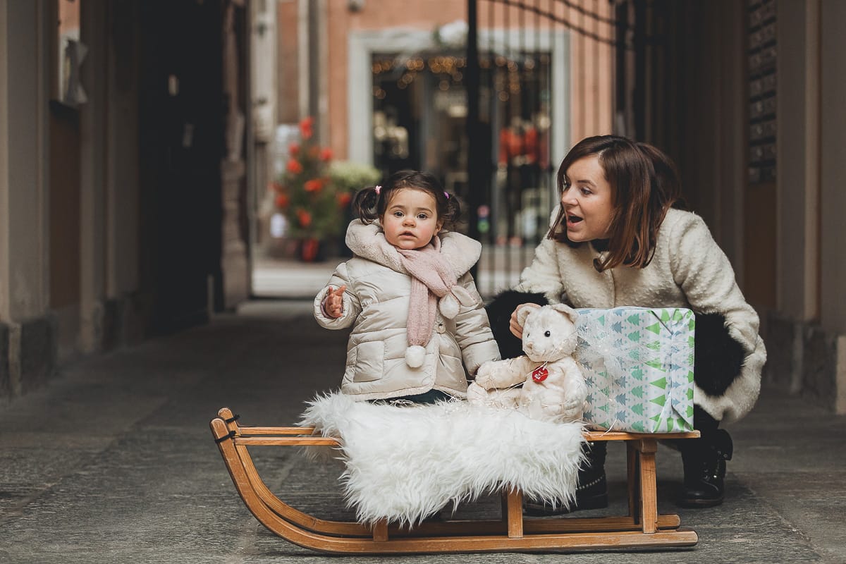 Fotografia di famiglia lifestyle di Natale. La piccola Lavinia con la sua mamma con un vecchio slittino di legno addobbato con un pellicciotto di lana di agnello bianco e pacchi regalo natalizi.