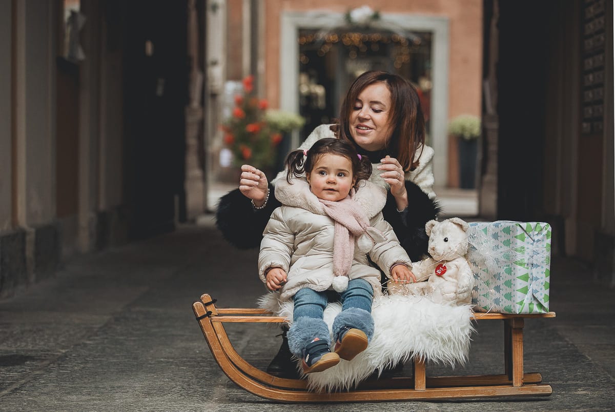 Fotografia di famiglia lifestyle di Natale. La piccola Lavinia con la sua mamma con un vecchio slittino di legno addobbato con un pellicciotto di lana di agnello bianco e pacchi regalo natalizi.