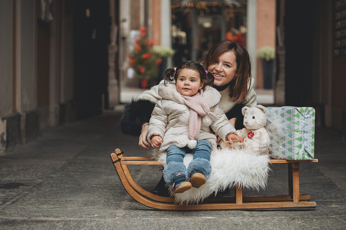 Fotografia di famiglia lifestyle di Natale. La piccola Lavinia con la sua mamma con un vecchio slittino di legno addobbato con un pellicciotto di lana di agnello bianco e pacchi regalo natalizi.