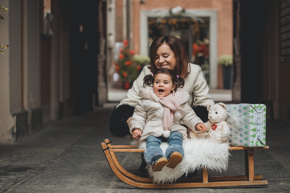 Fotografia di famiglia lifestyle di Natale. La piccola Lavinia con la sua mamma con un vecchio slittino di legno addobbato con un pellicciotto di lana di agnello bianco e pacchi regalo natalizi.