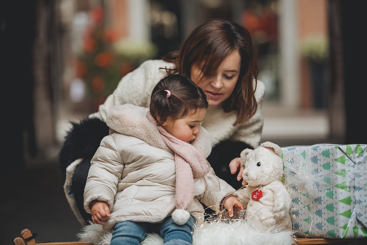 Fotografia di famiglia lifestyle di Natale. La piccola Lavinia con la sua mamma con un vecchio slittino di legno addobbato con un pellicciotto di lana di agnello bianco e pacchi regalo natalizi.