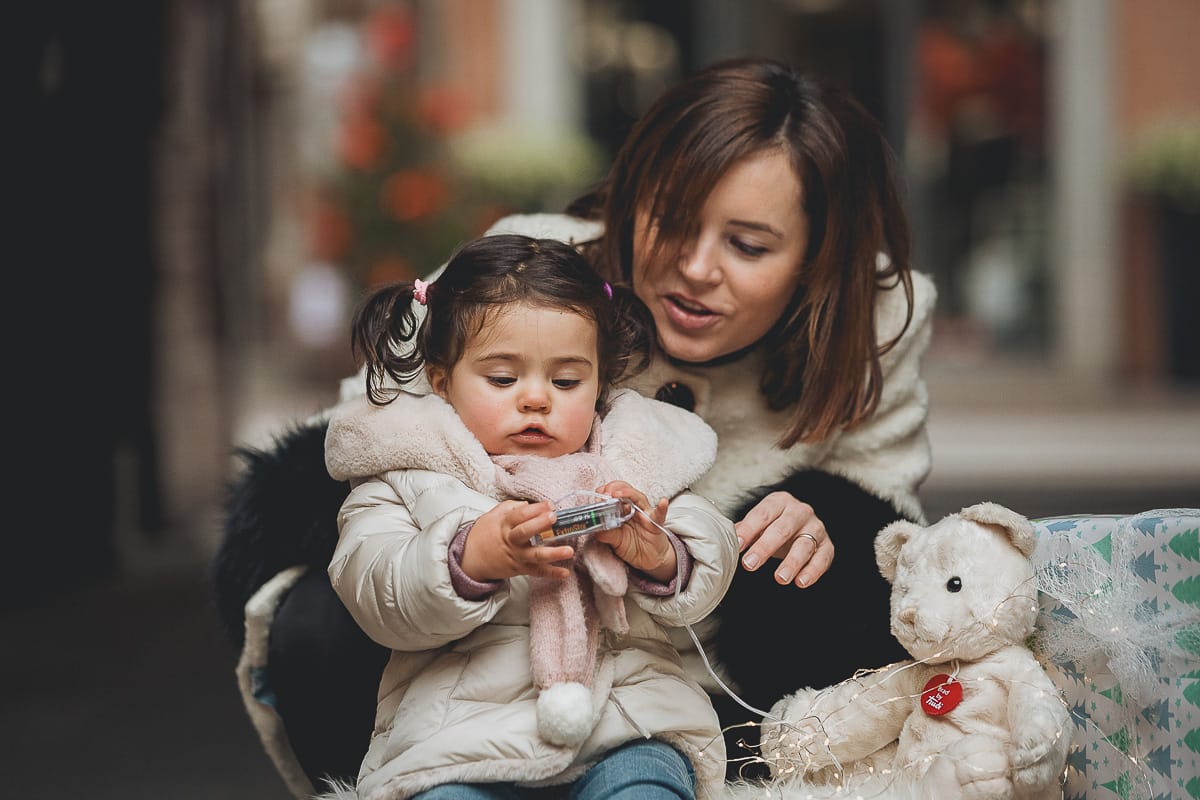 Fotografia di famiglia lifestyle di Natale. La piccola Lavinia con la sua mamma con un vecchio slittino di legno addobbato con un pellicciotto di lana di agnello bianco e pacchi regalo natalizi.