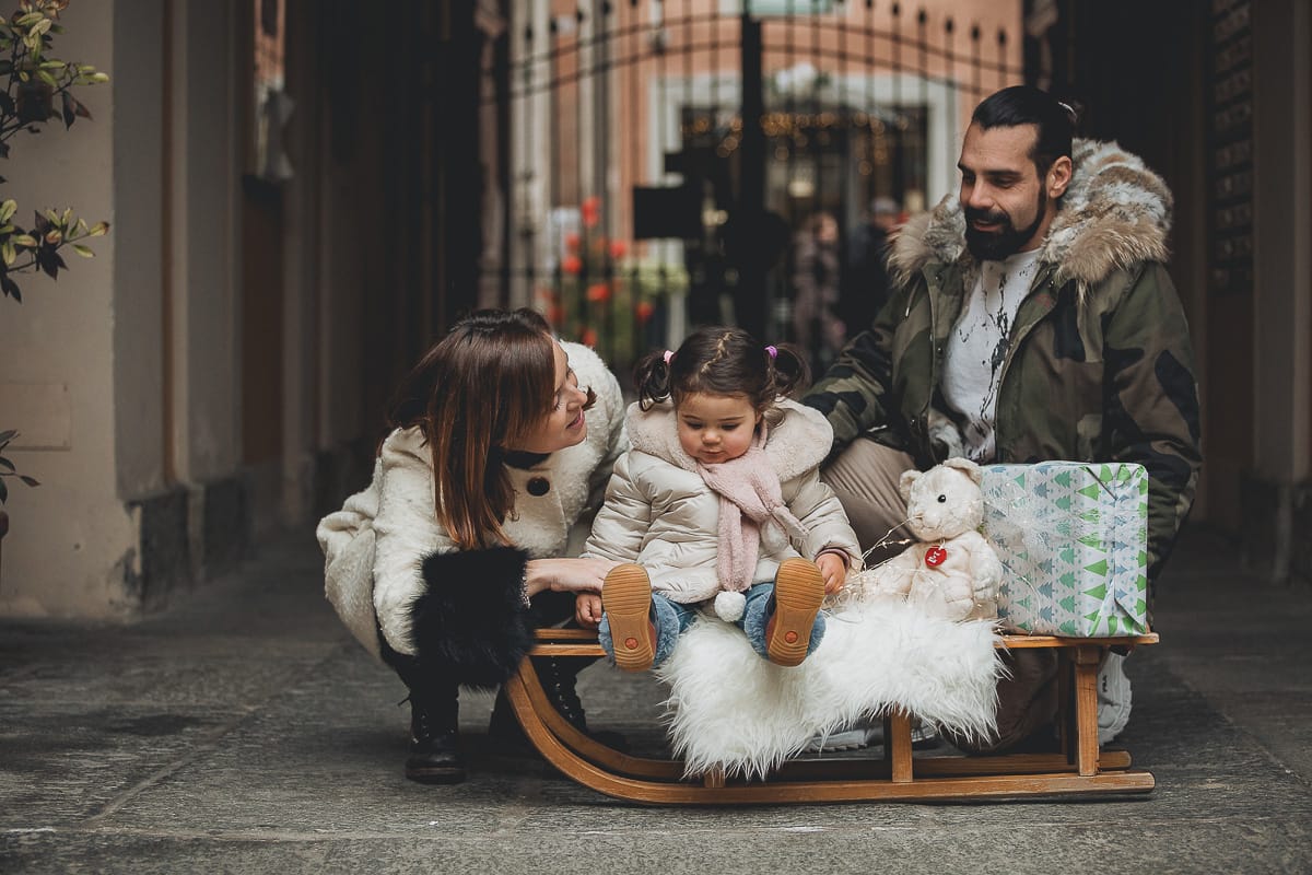 Fotografia di famiglia lifestyle di Natale. La piccola Lavinia con la sua mamma e il suo papà con un vecchio slittino di legno addobbato con un pellicciotto di lana di agnello bianco e pacchi regalo natalizi.