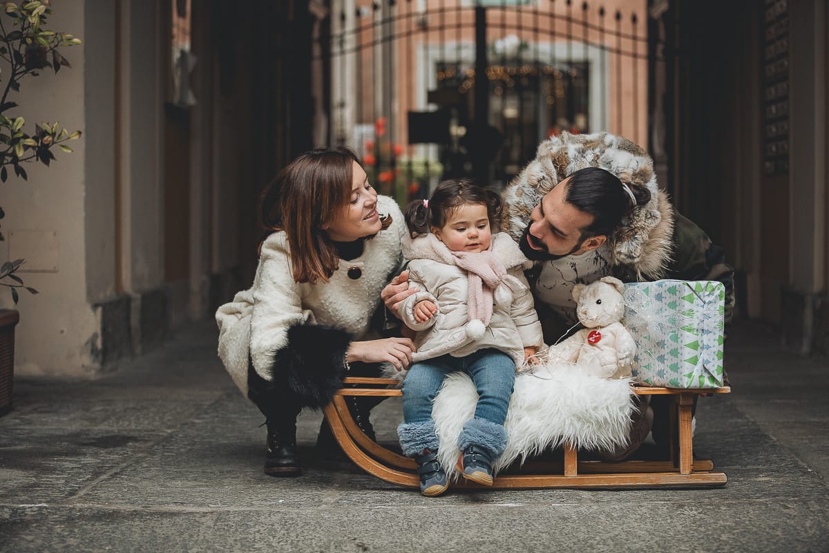Fotografia di famiglia lifestyle di Natale. La piccola Lavinia con la sua mamma e il suo papà con un vecchio slittino di legno addobbato con un pellicciotto di lana di agnello bianco e pacchi regalo natalizi.