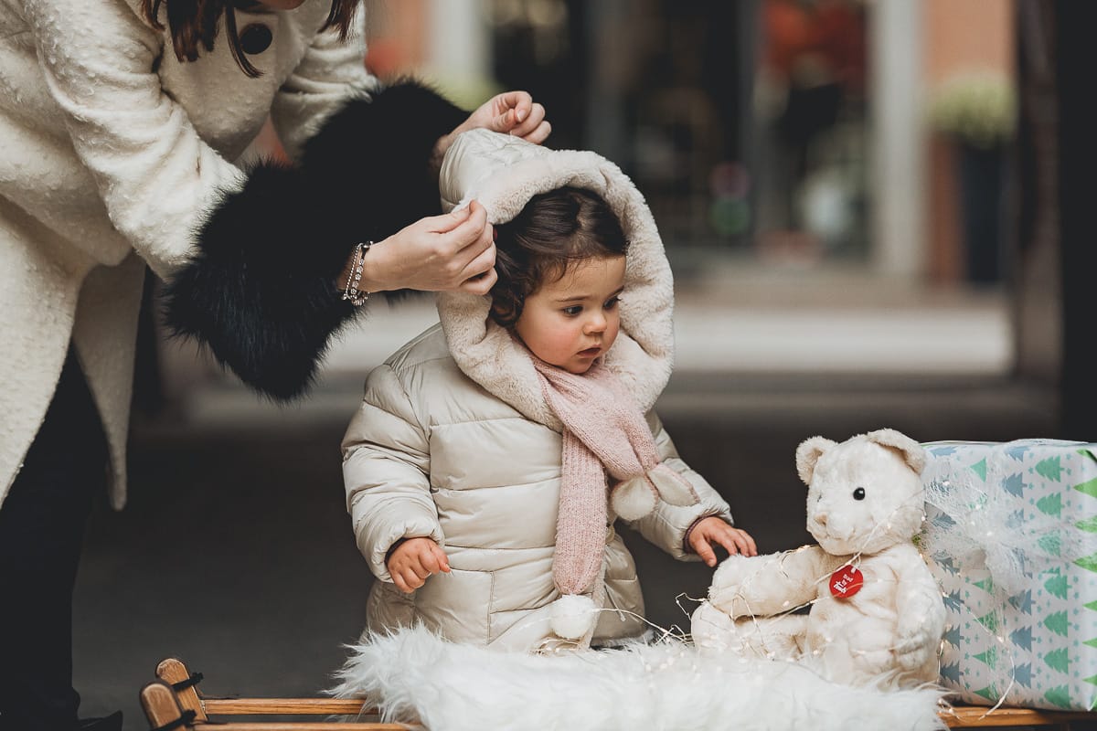 Fotografia di famiglia lifestyle di Natale. La piccola Lavinia con la sua mamma con un vecchio slittino di legno addobbato con un pellicciotto di lana di agnello bianco e pacchi regalo natalizi.