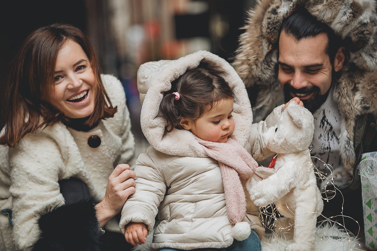 Fotografia di famiglia lifestyle di Natale. La piccola Lavinia con la sua mamma e il suo papà con un vecchio slittino di legno addobbato con un pellicciotto di lana di agnello bianco e pacchi regalo natalizi.