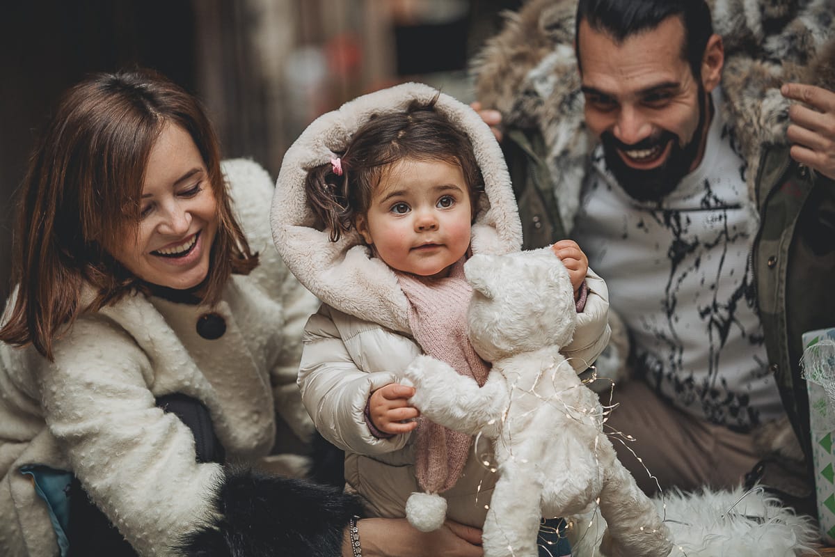 Fotografia di famiglia lifestyle di Natale. La piccola Lavinia con la sua mamma e il suo papà con un vecchio slittino di legno addobbato con un pellicciotto di lana di agnello bianco e pacchi regalo natalizi.