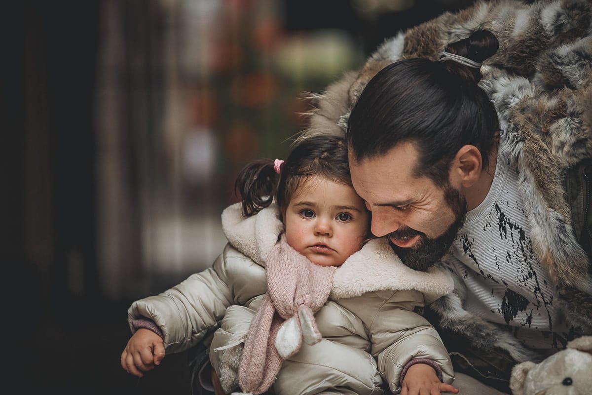 Fotografia di famiglia lifestyle di Natale. La piccola Lavinia con il suo papà con un vecchio slittino di legno addobbato con un pellicciotto di lana di agnello bianco e pacchi regalo natalizi.