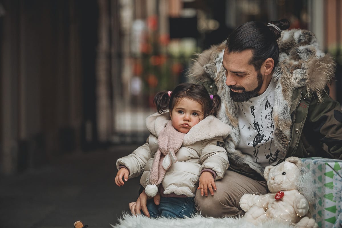 Fotografia di famiglia lifestyle di Natale. La piccola Lavinia con il suo papà con un vecchio slittino di legno addobbato con un pellicciotto di lana di agnello bianco e pacchi regalo natalizi.