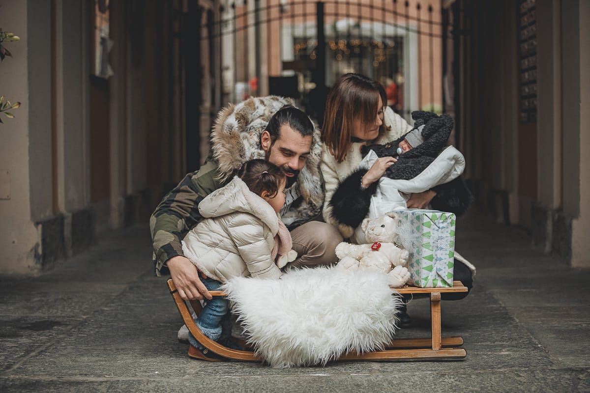 Fotografia di famiglia lifestyle di Natale. La piccola Lavinia con la sua mamma e il suo papà con un vecchio slittino di legno addobbato con un pellicciotto di lana di agnello bianco e pacchi regalo natalizi.