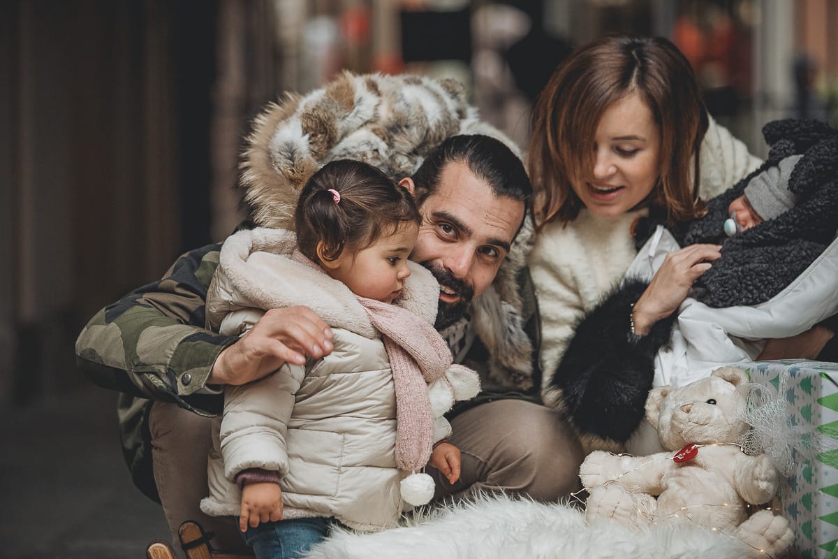 Fotografia di famiglia lifestyle di Natale. La piccola Lavinia con la sua mamma e il suo papà con un vecchio slittino di legno addobbato con un pellicciotto di lana di agnello bianco e pacchi regalo natalizi.