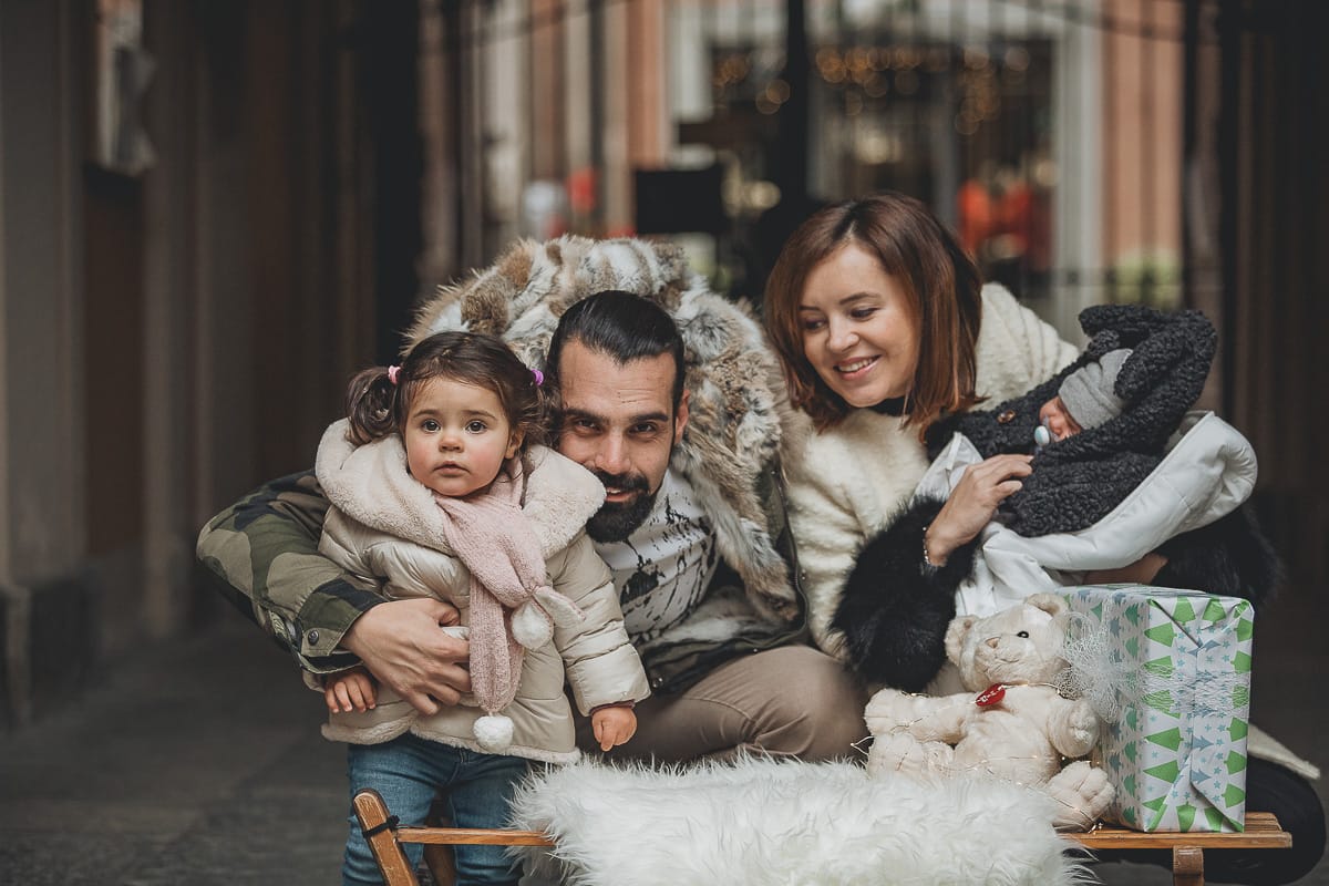 Fotografia di famiglia lifestyle di Natale. La piccola Lavinia con la sua mamma e il suo papà con un vecchio slittino di legno addobbato con un pellicciotto di lana di agnello bianco e pacchi regalo natalizi.