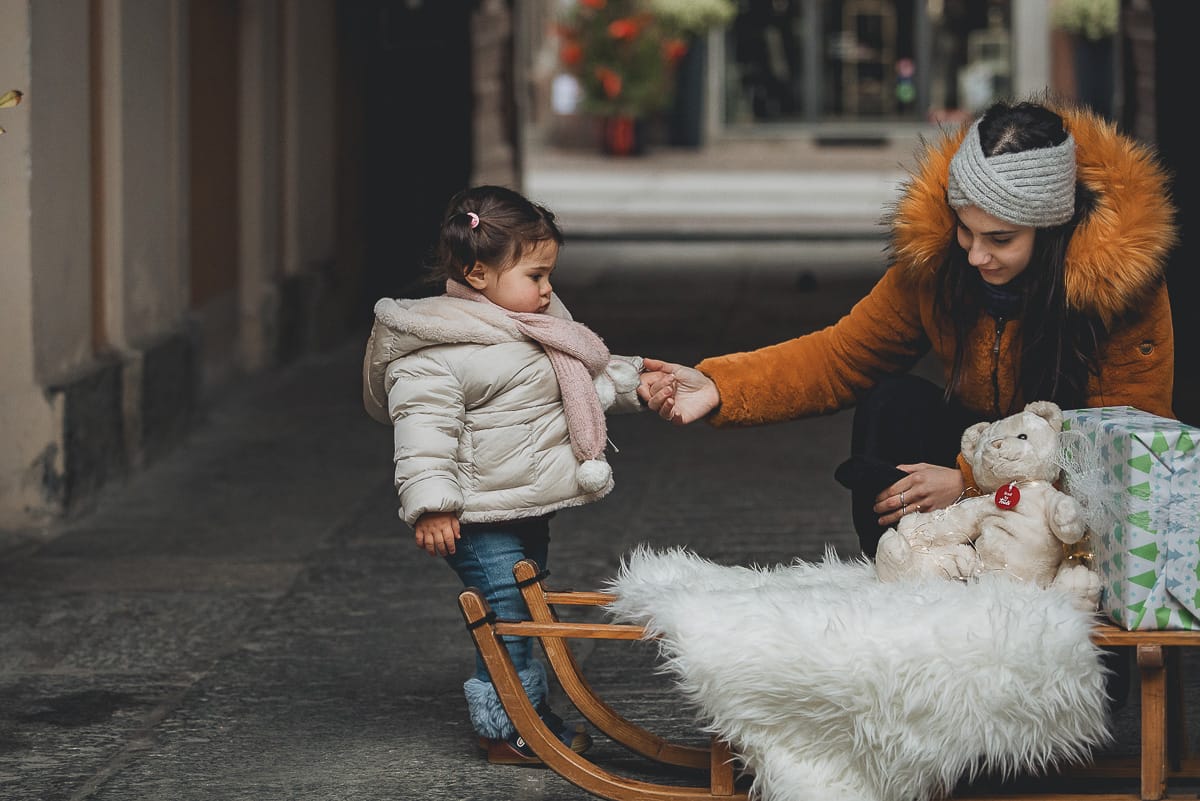 Fotografia di famiglia lifestyle di Natale. La piccola Lavinia con la zia e un vecchio slittino di legno addobbato con un pellicciotto di lana di agnello bianco e pacchi regalo natalizi.