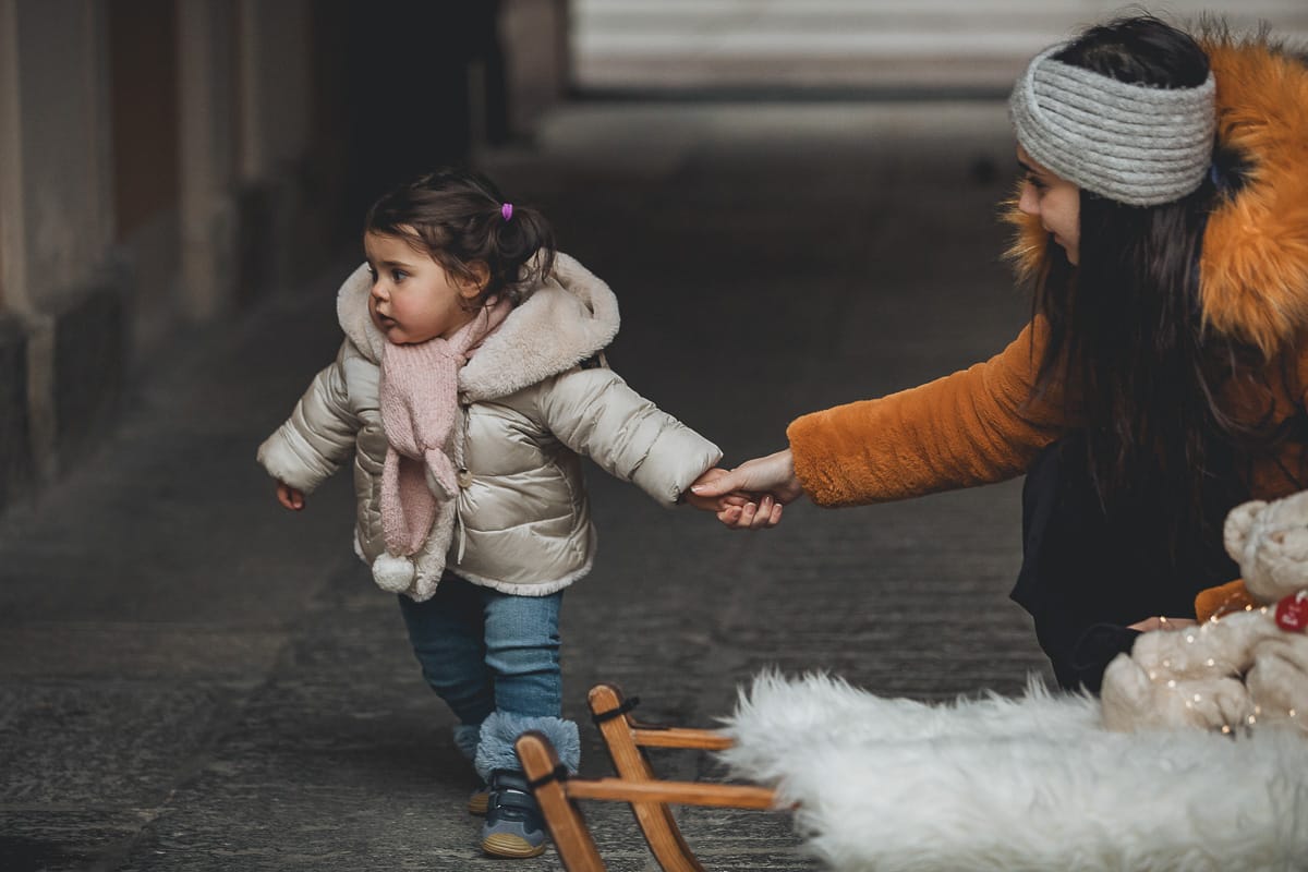 Fotografia di famiglia lifestyle di Natale. La piccola Lavinia con la zia e un vecchio slittino di legno addobbato con un pellicciotto di lana di agnello bianco e pacchi regalo natalizi.
