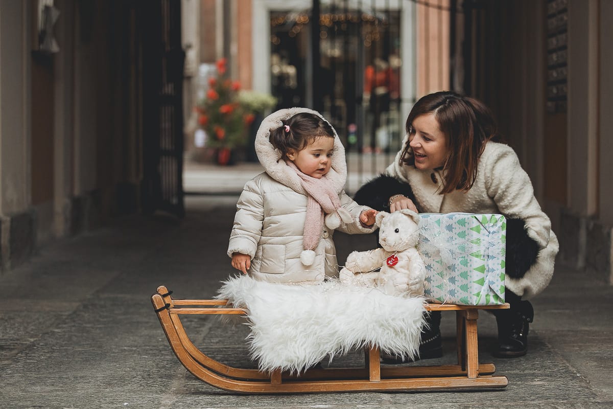 Fotografia di famiglia lifestyle di Natale. La piccola Lavinia con la sua mamma con un vecchio slittino di legno addobbato con un pellicciotto di lana di agnello bianco e pacchi regalo natalizi.
