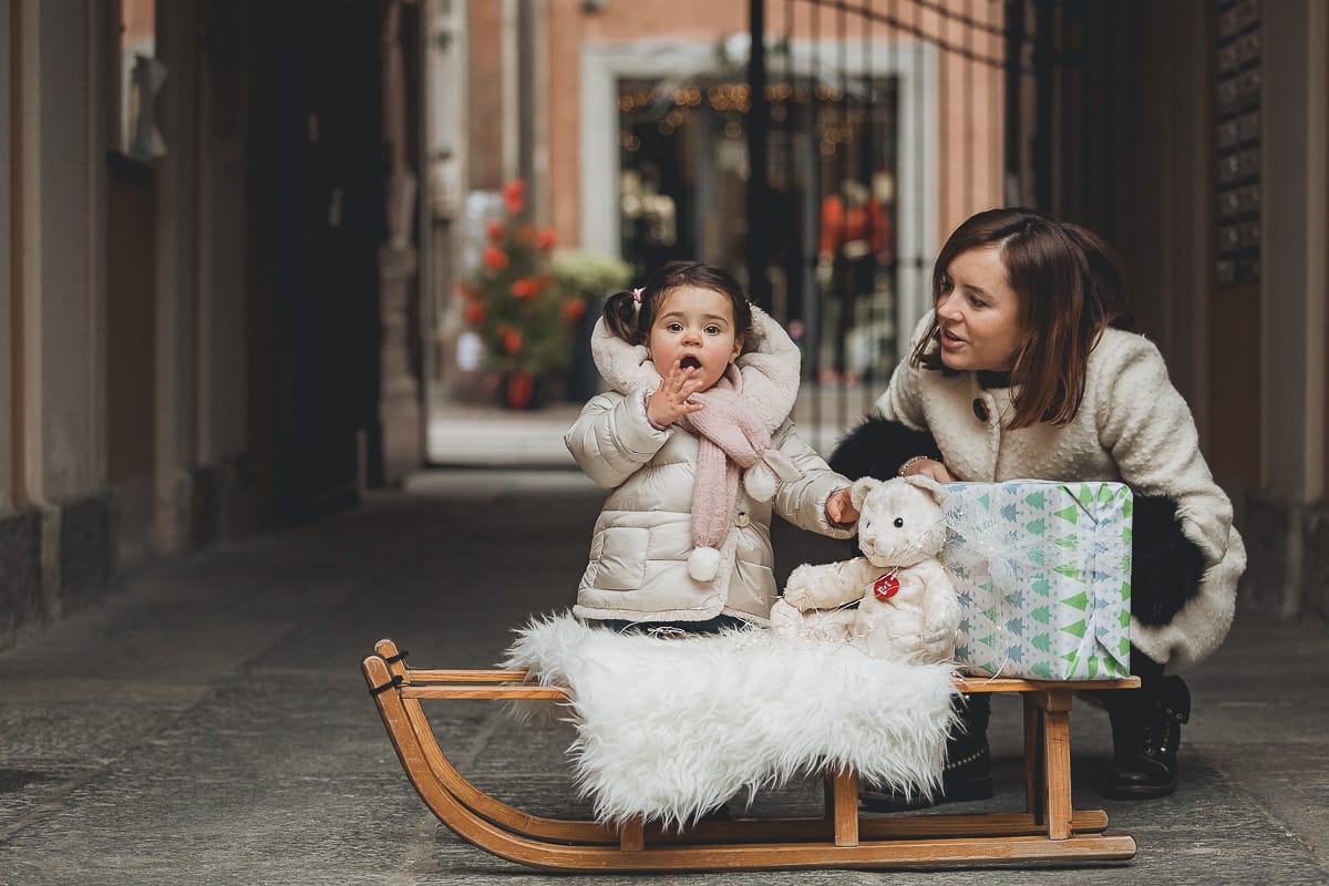 Fotografia di famiglia lifestyle di Natale. La piccola Lavinia con la sua mamma con un vecchio slittino di legno addobbato con un pellicciotto di lana di agnello bianco e pacchi regalo natalizi.