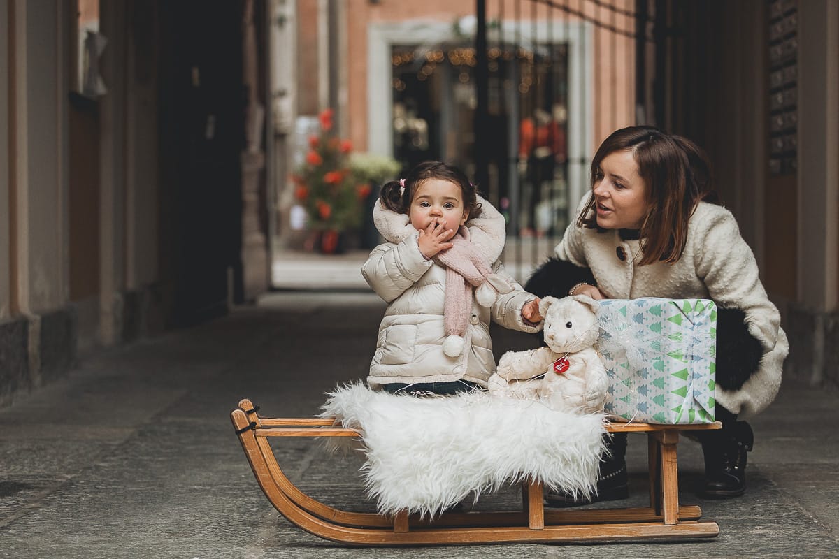 Fotografia di famiglia lifestyle di Natale. La piccola Lavinia con la sua mamma con un vecchio slittino di legno addobbato con un pellicciotto di lana di agnello bianco e pacchi regalo natalizi.