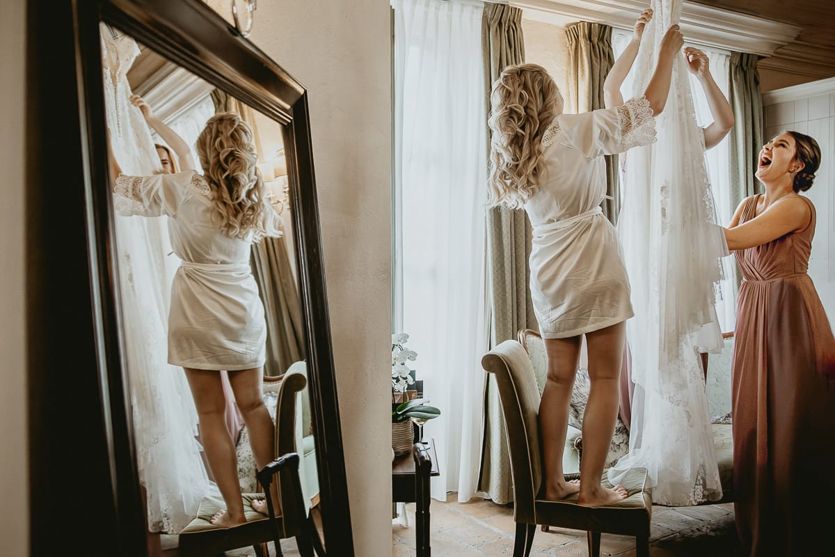 Destination wedding al Relais Sant'Uffizio. Preparazione della sposa. Sposa che prende il suo abito, riflessa nello specchio con una damigella che la aiuta.