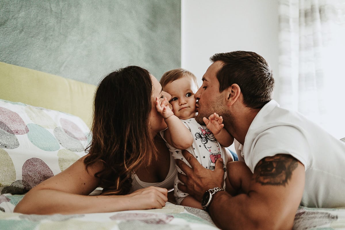 Fotografia di famiglia a Chieri. La piccola Aurora sul lettone con i genitoriche la baciano