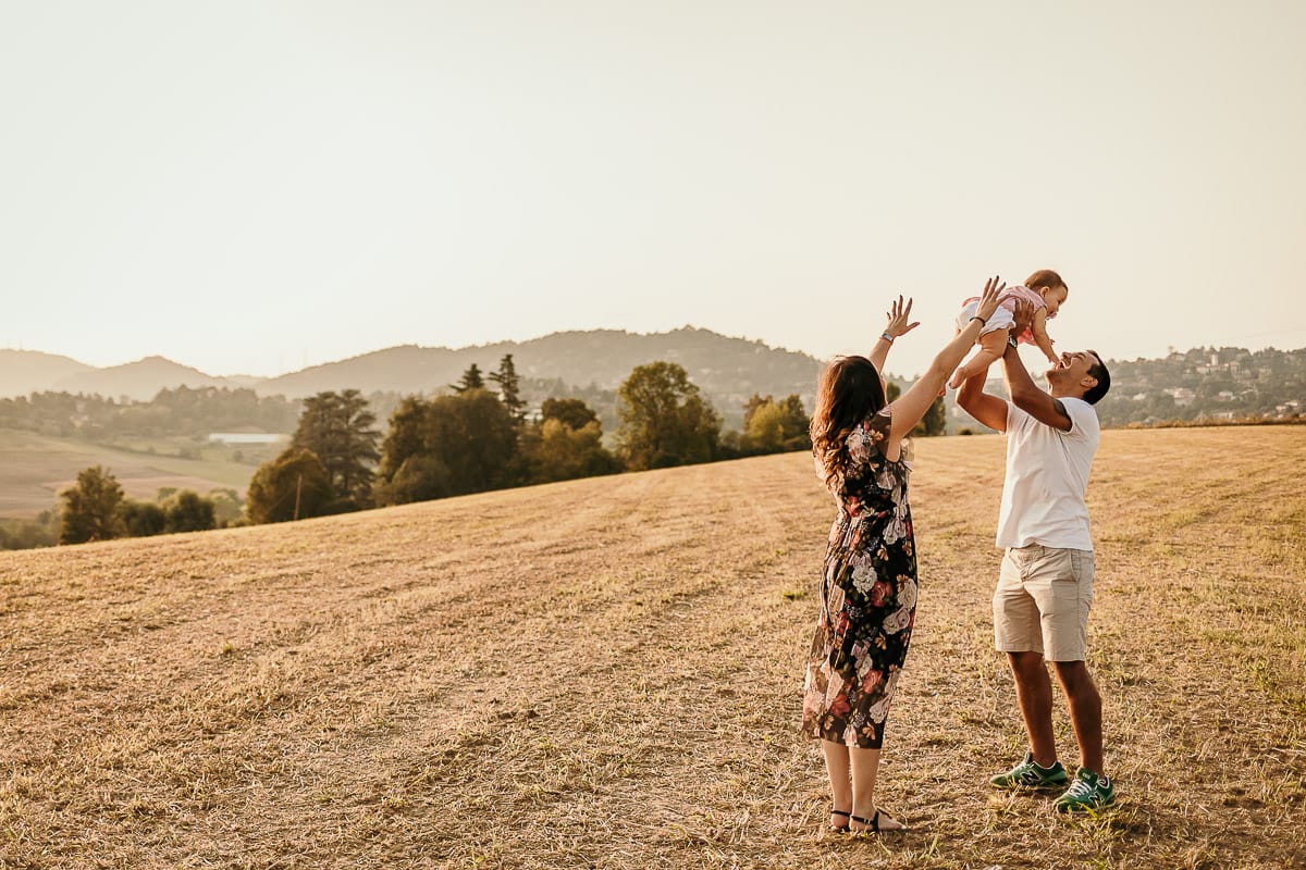 Fotografia di famiglia a Chieri. Ritratti spontanei in esterno nelle colline intorno alla città, al tramonto. Silhouette del papà che fa volare la bambina in controluce e la mamma protende le braccia.